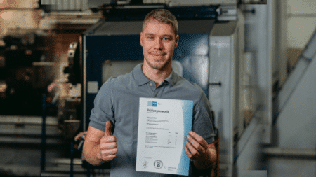 Ein Mann in einem grauen Poloshirt steht drinnen, lächelt und hält in seiner rechten Hand ein Zertifikat, während er mit seiner linken Hand den Daumen hoch zeigt. Im Hintergrund sind Industriemaschinen zur Herstellung von Schmiedeteilen zu sehen.