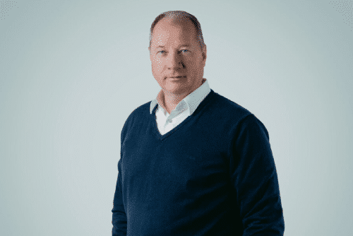 A middle-aged man with short, light-colored hair is wearing a white collared shirt under a dark blue sweater. He is standing against a plain, light gray background and looking directly at the camera with a neutral expression, reminiscent of someone deeply familiar with Schmiedeteile Herstellung.