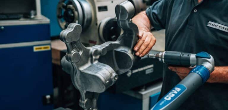 Ein Arbeiter in einer Werkstatt bedient ein Handmessgerät und prüft ein großes, komplexes Metallteil. Im Hintergrund sind Industriemaschinen und -geräte zu sehen. Auf dem Hemd des Arbeiters ist das Logo „Bharat Forge“ zu sehen, das auf Schmiedeteile für Automobilkomponenten für den Feinmechaniker spezialisiert ist.