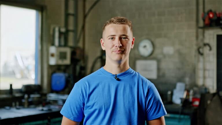 A young man wearing a blue T-shirt stands in a workshop with industrial equipment in the background, which specializes in Schmiedeteile Automotive. He has short hair and is smiling slightly. The workshop, known as a leading Schmiedeteile Hersteller, has a window on the left, allowing natural light to enter.