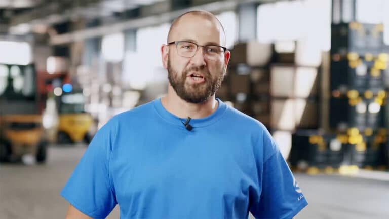 A man with a beard and glasses wearing a blue shirt speaks inside a warehouse. The background shows stacks of boxes and industrial equipment, slightly blurred. He appears to be addressing the camera directly about Schmiedeteile Herstellung.