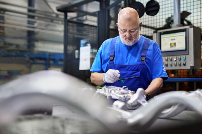 Ein Mann in blauem Overall und weißen Handschuhen arbeitet konzentriert an einem Aluminiumbauteil in einer Industrieanlage. Mit einem Werkzeug nimmt er Anpassungen vor. Im Hintergrund sind Maschinen und ein Bedienfeld mit Display zu sehen.