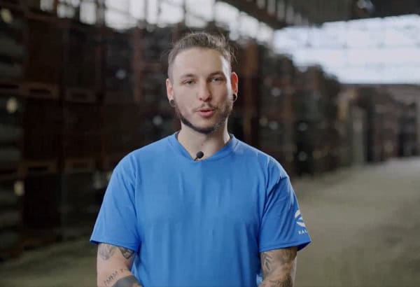 Eine tätowierte Person in einem blauen Hemd spricht in ein Mikrofon in einer industriellen Umgebung. Der Hintergrund, gefüllt mit Metallstapeln und -regalen, lässt auf ein Lager schließen, das auf Aluminium-Schmiedeteile oder ähnliche Komponenten spezialisiert ist.