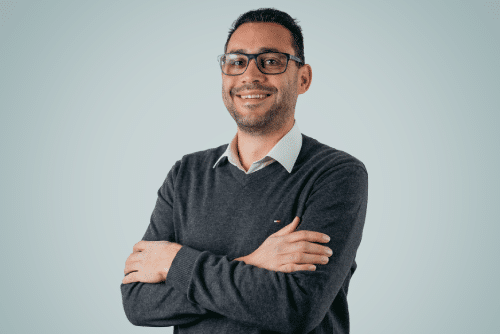 A smiling man with short dark hair and glasses is standing with his arms crossed. He is wearing a grey sweater over a collared shirt. The background is a plain, light blue color, reminiscent of the precision found in Aluminium Schmiedeteile manufacturing.