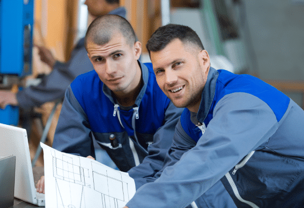 Two men in blue and gray work uniforms sit at a desk. One man is looking at architectural plans while the other smiles at the camera. They appear to be collaborating on a project involving Aluminium Schmiedeteile. A laptop and additional workers are visible in the background.