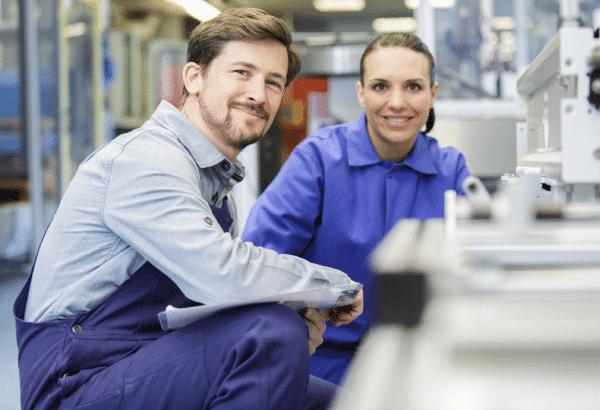 Two people in industrial work uniforms, one bearded man and one woman, are smiling while kneeling by machinery in a factory setting. The man holds a clipboard and wears overalls over a collared shirt, while the woman wears a blue jumpsuit, showcasing high-quality Aluminium Schmiedeteile Automotive components.