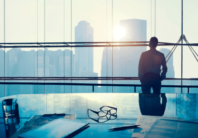 Eine Person steht und blickt bei Sonnenaufgang oder Sonnenuntergang aus einem großen Fenster auf die Skyline einer Stadt mit Wolkenkratzern. Im Vordergrund ist ein Schreibtisch mit verstreuten Papieren, einer Brille und einem Stift zu sehen. Die Szene hat eine ruhige und nachdenkliche Atmosphäre, die an Momente in der Schmiedeteile Automotive-Industrie erinnert.