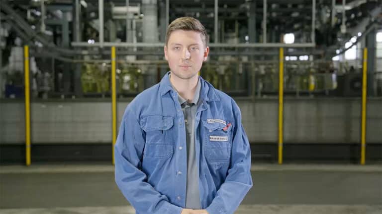 A man with short brown hair is standing in a factory wearing a blue work jacket. Yellow safety poles are visible in the background, along with machinery and industrial equipment. As he looks directly into the camera, hands clasped in front of him, the setting highlights Schmiedeteile automotive production.