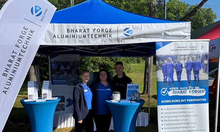 Eine Gruppe von drei Personen steht vor einem blau-weißen Zelt und Bannern mit dem Logo von „Bharat Forge Aluminiumtechnik“. Der Aufbau mit zwei hohen blauen runden Tischen mit Informationsmaterial unterstreicht ihre Kompetenz als Schmiedeteilehersteller, und das alles vor einer sonnigen Außenkulisse.