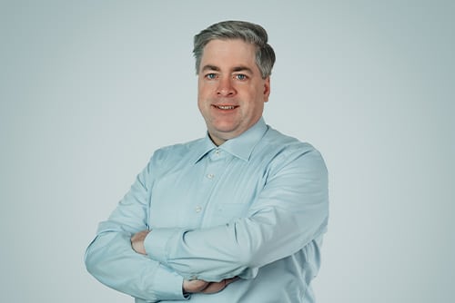 A man with short gray hair is wearing a light blue button-down shirt. He is smiling and has his arms crossed over his chest, exuding confidence like an expert in Aluminium Schmiedeteile Herstellung. The background is plain and light-colored.