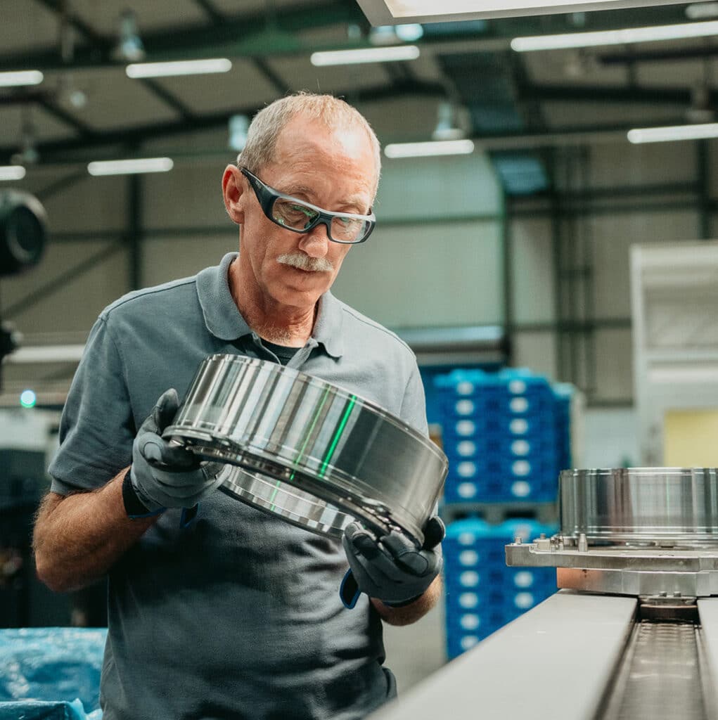Ein älterer Mann mit Schutzbrille und Handschuhen prüft ein großes Metallteil in einer industriellen Fabrikumgebung mit Regalen und Maschinen im Hintergrund.