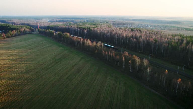 Aerial view of a long train passing through a forested area bordered by a large field. The field has a mix of green and brown patches, reminiscent of the varied texture of Schmiedeteile. The sky is clear, and there is a small town visible in the distant background.