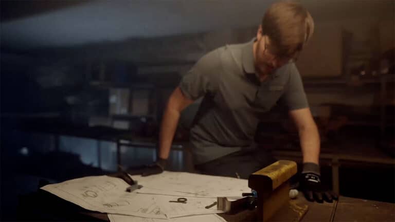 A person wearing safety gloves works in a dimly lit workshop. The individual is hunched over a table cluttered with blueprints and tools, focusing intently on the task at hand. The room appears industrial, typical of a Schmiedeteile Hersteller, with various equipment in the background.