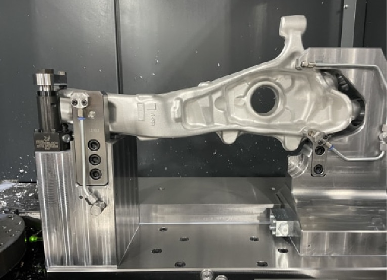 A metallic automotive suspension part is secured in a custom fixture on a workbench, likely in a manufacturing or inspection facility. The background is comprised of gray metal panels.