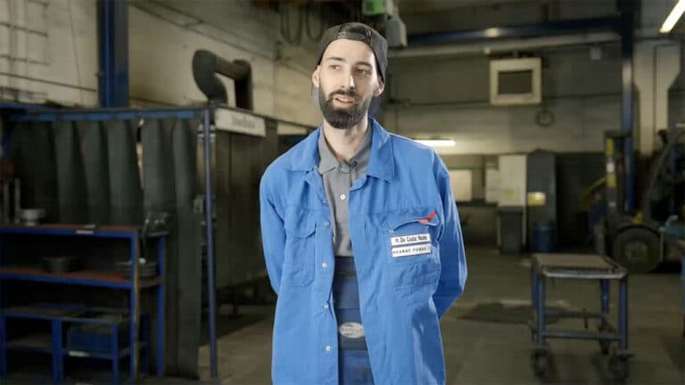 A person with a beard and mustache, wearing a blue work jacket and a baseball cap backward, stands in an industrial workshop. The space, filled with various equipment, workbenches, and machinery for Schmiedeteile Herstellung, suggests an active setting where the individual may be explaining something.