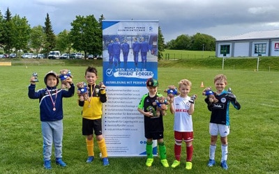 Eine Gruppe von sechs kleinen Kindern steht auf einer Wiese, hält Trophäen in den Händen und lächelt in die Kamera. Sie tragen verschiedene Fußballtrikots. Hinter ihnen hängt ein Banner mit Text und Bildern über Schmiedeteile Herstellung und im Hintergrund sind Bäume und Gebäude zu sehen.