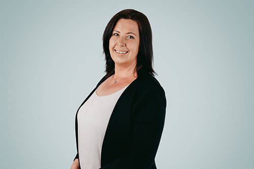 A person with shoulder-length dark hair, wearing a black blazer over a white top, smiles while standing against a light blue background, embodying the professionalism of a Schmiedeteile Hersteller.