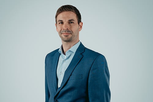 A man with short hair is standing against a light blue background. He is wearing a blue blazer over a white shirt and is looking directly at the camera with a neutral expression, embodying the precision and quality often associated with Aluminium Schmiedeteile.