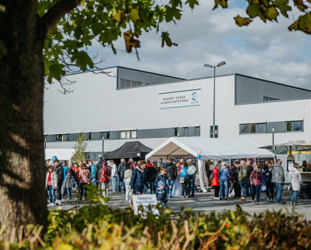 Eine Menschenmenge versammelt sich draußen in der Nähe eines großen, weißen Industriegebäudes mit der Aufschrift „Diehl Aviation“ unter blauem Himmel. Man sieht Zelte und Stände, einige Leute halten Luftballons in den Händen. Grüne, belaubte Zweige rahmen den oberen Bildrand ein und vermitteln ein Gemeinschaftsgefühl rund um diesen Schmiedeteile-Hersteller.