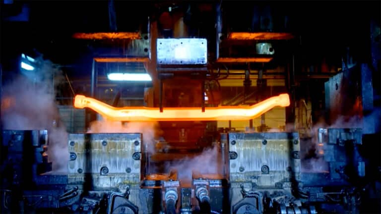 A close-up of a factory machine processing a glowing, heated, orange metal piece. Steam surrounds the equipment as the metal is being shaped or forged. Industrial elements and other machinery parts are visible in the background, bathed in blue and orange light, highlighting Schmiedeteile Herstellung.