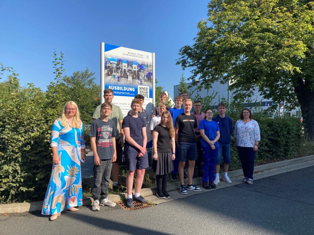 Eine Gruppe von Menschen posiert im Freien vor einem Schild mit der Aufschrift „Ausbildung“. Die Gruppe, möglicherweise zukünftige Schmiedeteilehersteller, besteht aus Personen unterschiedlichen Alters, die auf einer gepflasterten Fläche mit Grünflächen und Bäumen im Hintergrund stehen. Der Himmel ist klar und blau.