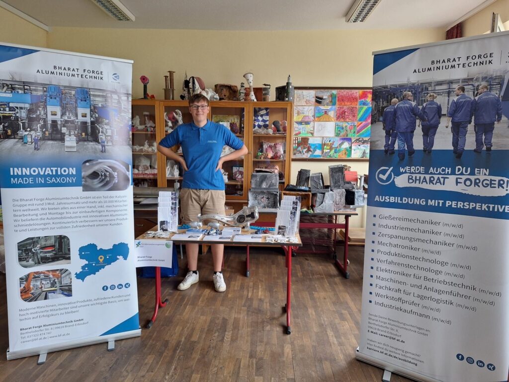 A person stands behind a table with displayed mechanical parts and tools at an exhibition booth. The booth features two large banners, highlighting "Innovation Made in Saxony" and recruitment for Bharat Forge Aluminumtechnik. The background shows colorful artwork.