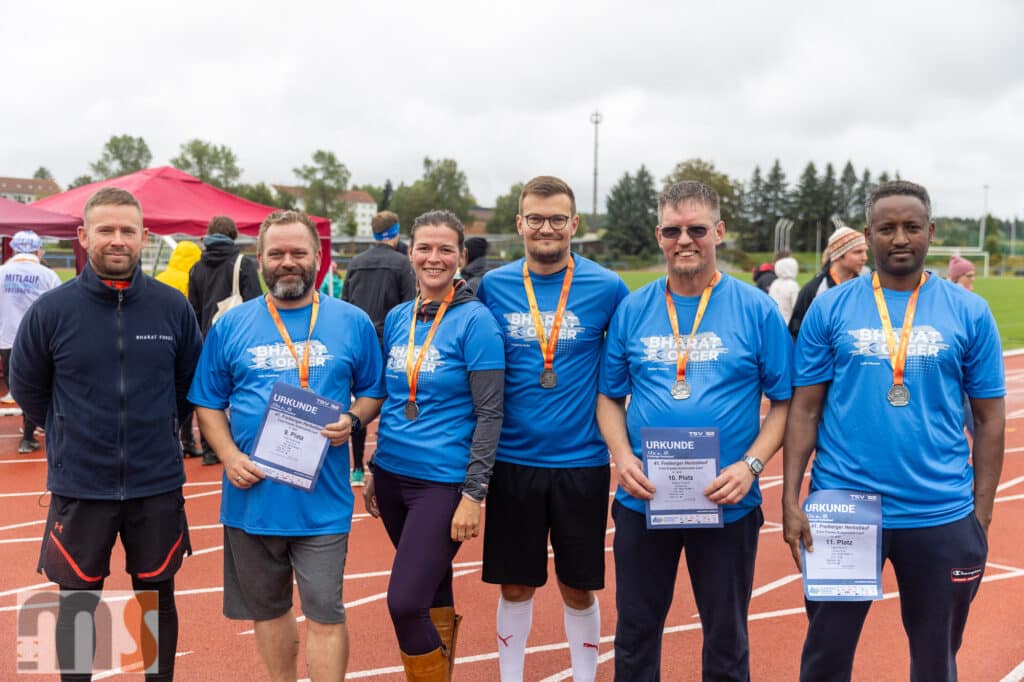 Fünf Menschen stehen auf einer Freiluftstrecke. Sie tragen blaue T-Shirts und Medaillen um den Hals. Sie halten Zertifikate in den Händen und lächeln. Im Hintergrund sind ein Zelt und weitere Menschen zu sehen. Der Himmel ist bedeckt.