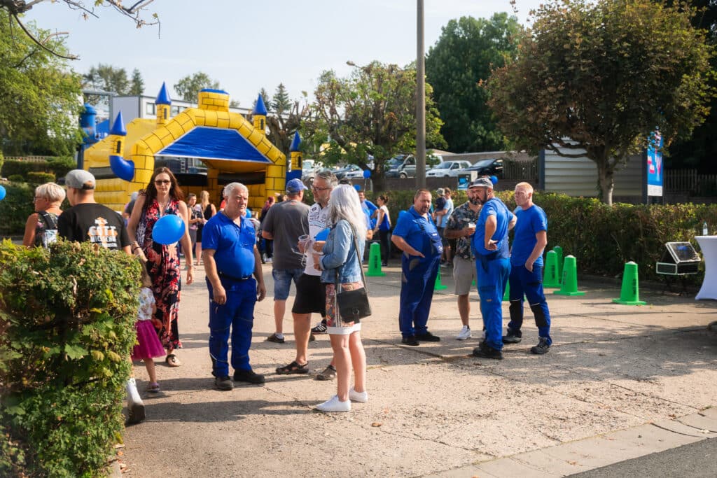 A group of people gathered outdoors near a large yellow and blue inflatable structure. Some people are wearing blue uniforms, while others are in casual clothing. Green traffic cones and trees are visible in the background. A man holds a blue balloon.