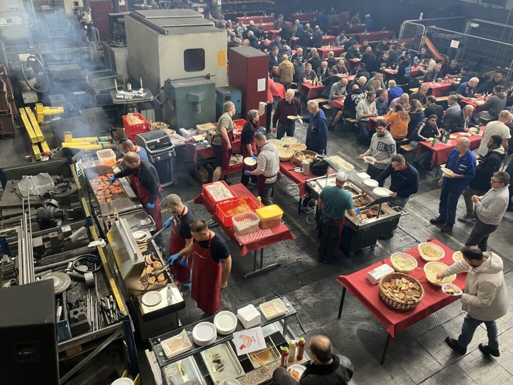 Eine große Gruppe von Menschen hat sich in einem Industriegebäude versammelt und genießt eine gemeinsame Mahlzeit. Mehrere Tische sind mit Essen aufgestellt und mehrere Menschen arbeiten an Kochstationen. Die Atmosphäre ist geschäftig und lebhaft, die Menschen sitzen, essen und unterhalten sich.