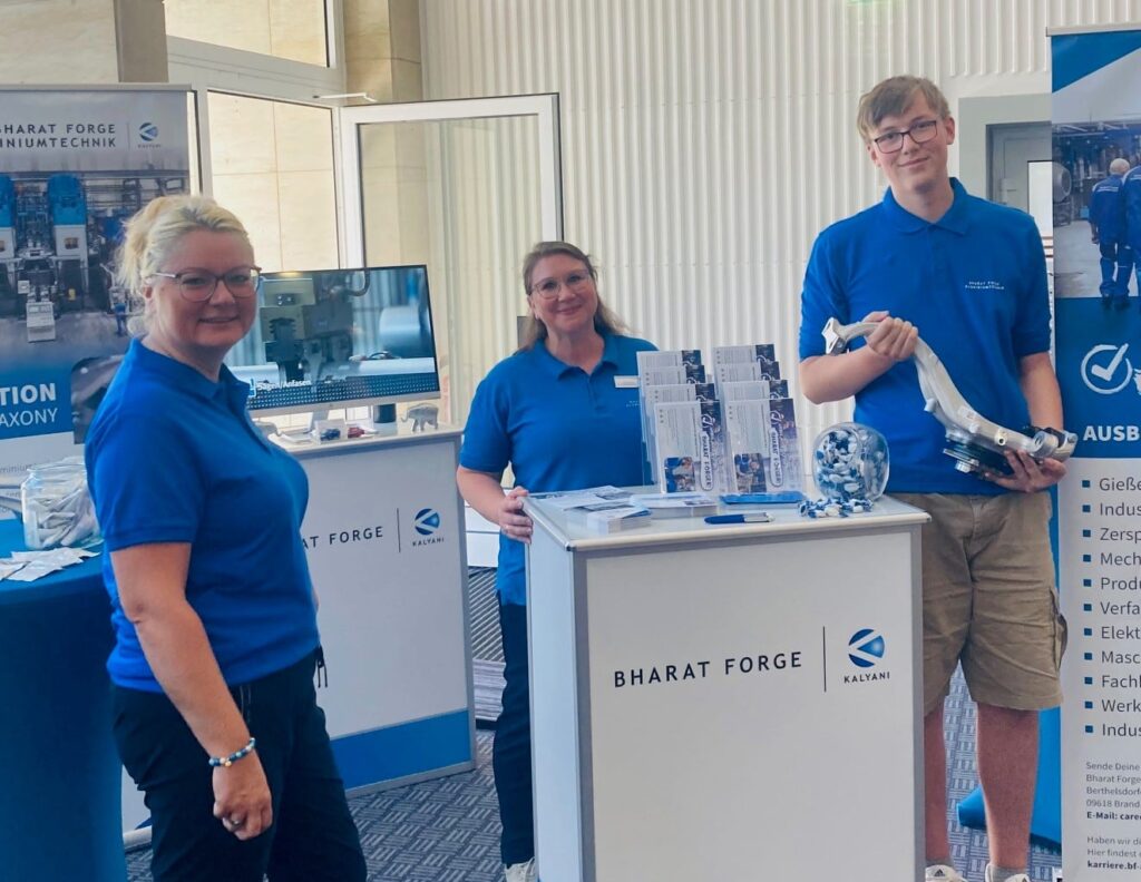 Three people in blue shirts stand around a promotional booth for Bharat Forge. Two women stand on the left and behind the booth, while a young man on the right holds a metallic object. The booth features pamphlets, glassware, and a banner in the background.