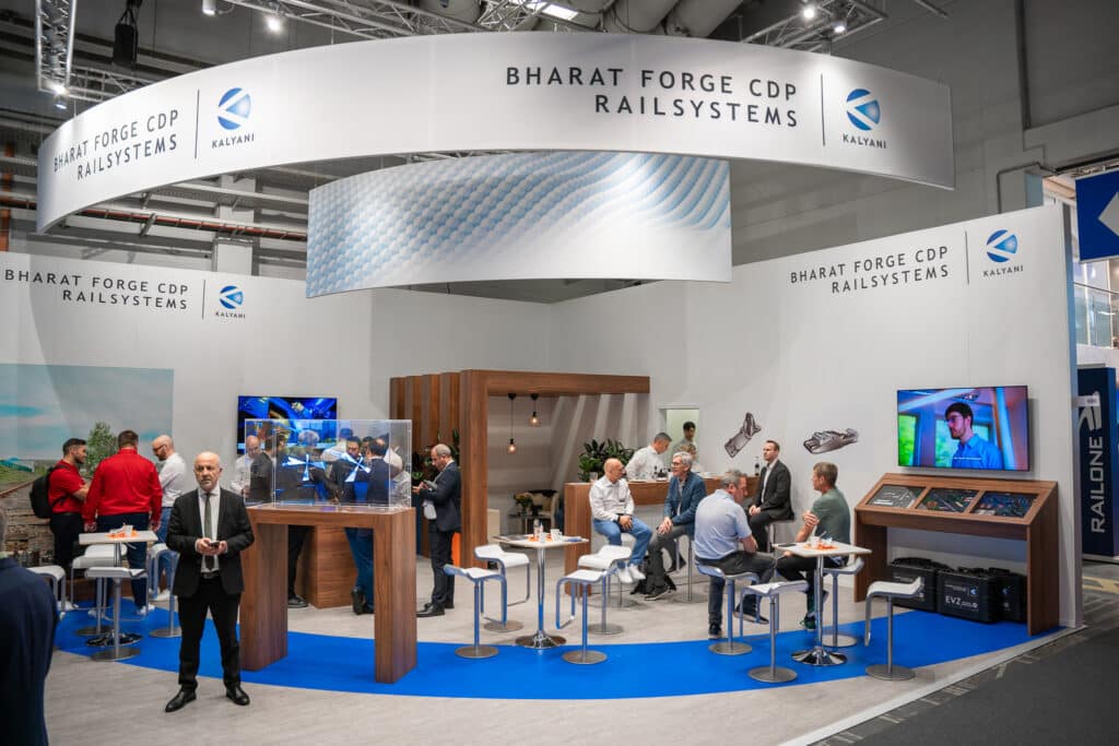 Messestand für Bharat Forge CDP Rail Systems mit Menschen, die miteinander interagieren. Der Stand verfügt über moderne Displays, einen Bildschirm mit Videos und eine Glasvitrine mit Teilen. Farbschema in Weiß und Blau mit prominenter Darstellung von Bharat Forge und zugehöriger Marke.