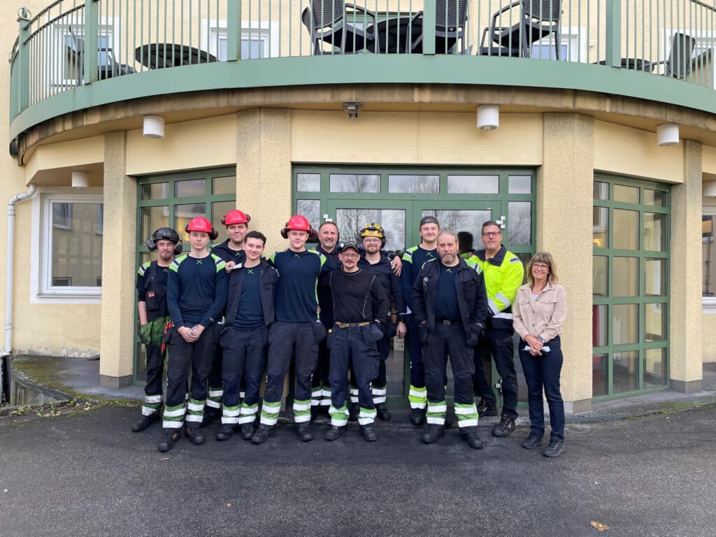 Eine Gruppe von zehn Feuerwehrmännern in Uniform und Helm steht vor einem Gebäude mit großen Fenstern. Zwei Personen in Zivil stehen auf beiden Seiten der Gruppe. Sie lächeln alle.