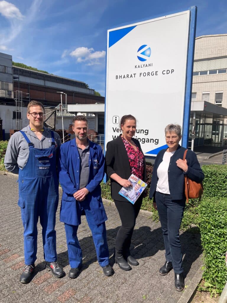 Vier Personen stehen an einem sonnigen Tag vor einem Schild mit der Aufschrift „Kalyani Bharat Forge CDP“. Zwei tragen blaue Arbeitsuniformen, die anderen beiden sind in Business-Kleidung gekleidet. Im Hintergrund sind Industriegebäude zu sehen.