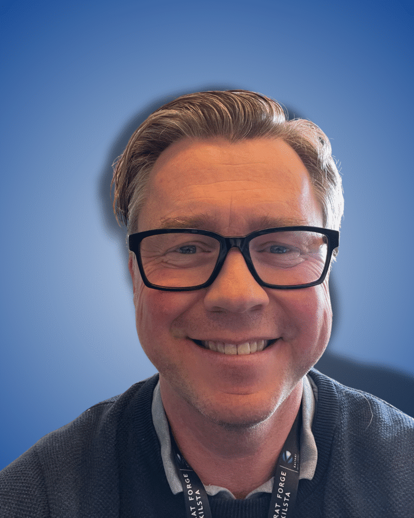 Ein Mann mit hellbraunem Haar und schwarzer Brille lächelt vor einem blau abgestuften Hintergrund in die Kamera. Er trägt einen dunklen Pullover und ein Schlüsselband.