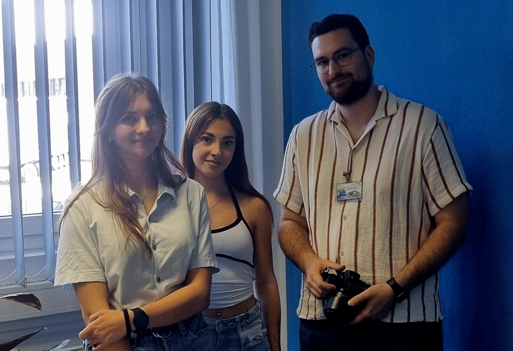 Drei Personen stehen in einem Innenraum vor einem Fenster mit vertikalen Jalousien. Zwei Frauen auf der linken Seite, beide lächelnd, und ein Mann mit Brille und Bart auf der rechten Seite, der eine Kamera hält. Die Wand hinter ihnen ist blau.
