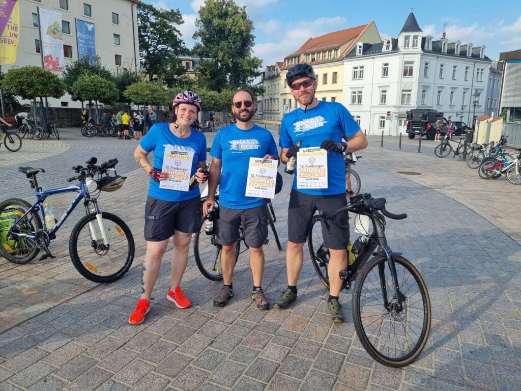 Drei Radfahrer in blauen Hemden stehen mit ihren Fahrrädern auf einem kopfsteingepflasterten Platz, lächeln und halten Urkunden in der Hand. Im Hintergrund sind in der Nähe von Gebäuden einige weitere Menschen und Fahrräder zu sehen.