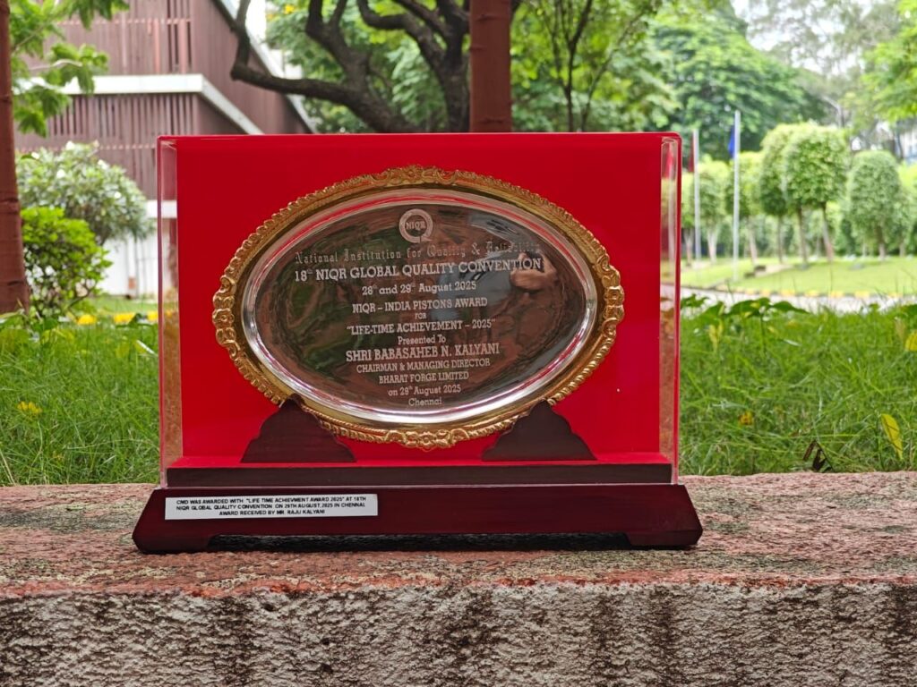 Eine silberne und goldene Plakette auf einem roten Sockel steht auf einem Steinsockel im Freien, umgeben von Grünpflanzen. Die Plakette erinnert an die 16. NIGQ Global Quality Convention und wird Shri Basavaraj H. Kavatagi überreicht.