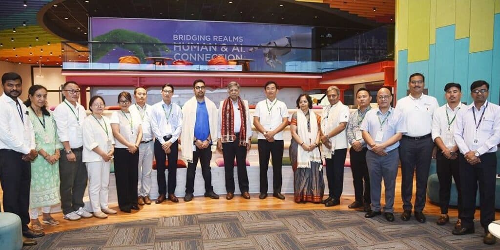 Eine Gruppe von 17 Männern und Frauen steht in einer Reihe und posiert für ein Foto in einem farbenfrohen, modernen Gebäude mit einem Schild, auf dem "Bridging Realms: Human & AI Connection" im Hintergrund.