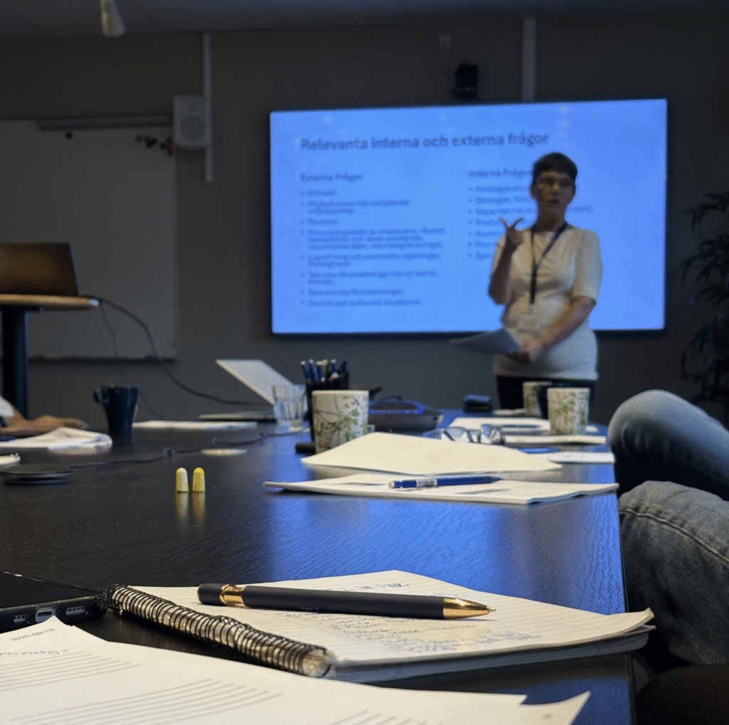 Eine Frau steht vor einem Bildschirm, auf dem ein schwedischer Text angezeigt wird, und spricht zu einer Gruppe, die während einer Sitzung oder eines Workshops an einem Tisch mit Notizbüchern, Stiften und Tassen sitzt.