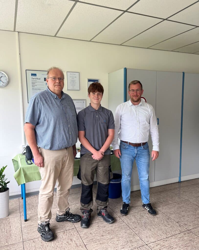 Drei Personen stehen in einem Innenraum auf einem gefliesten Boden und posieren für ein Gruppenfoto vor einem Schrank und einem grün gedeckten Tisch. Sie sind leger gekleidet, und an der Wand hinter ihnen sind Zertifikate zu sehen.