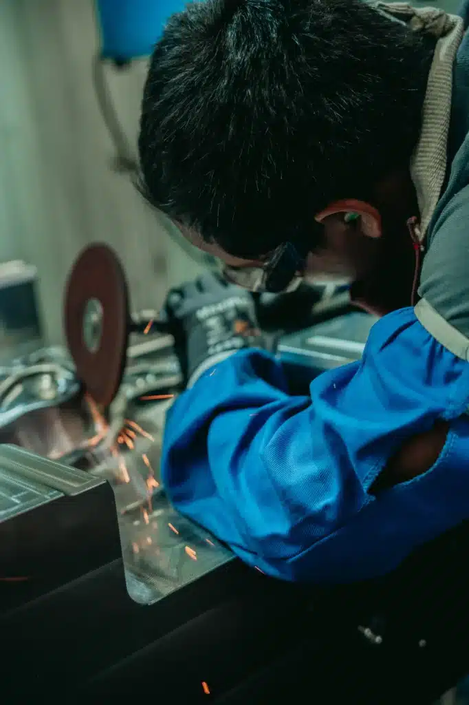 A person wearing safety glasses and a blue protective jacket uses a grinder to cut metal, producing sparks in a workshop setting.