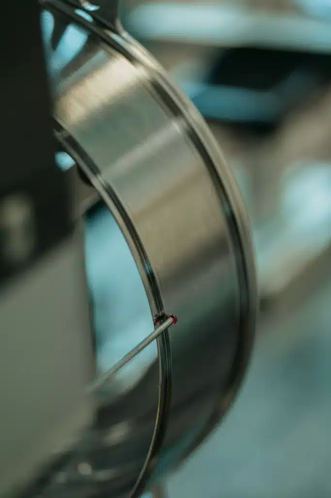 A close-up of a metallic spool or coil, with a thin, red-tipped wire being guided along its edge. The background is blurred, drawing focus to the shiny surface and the detail of the wire.