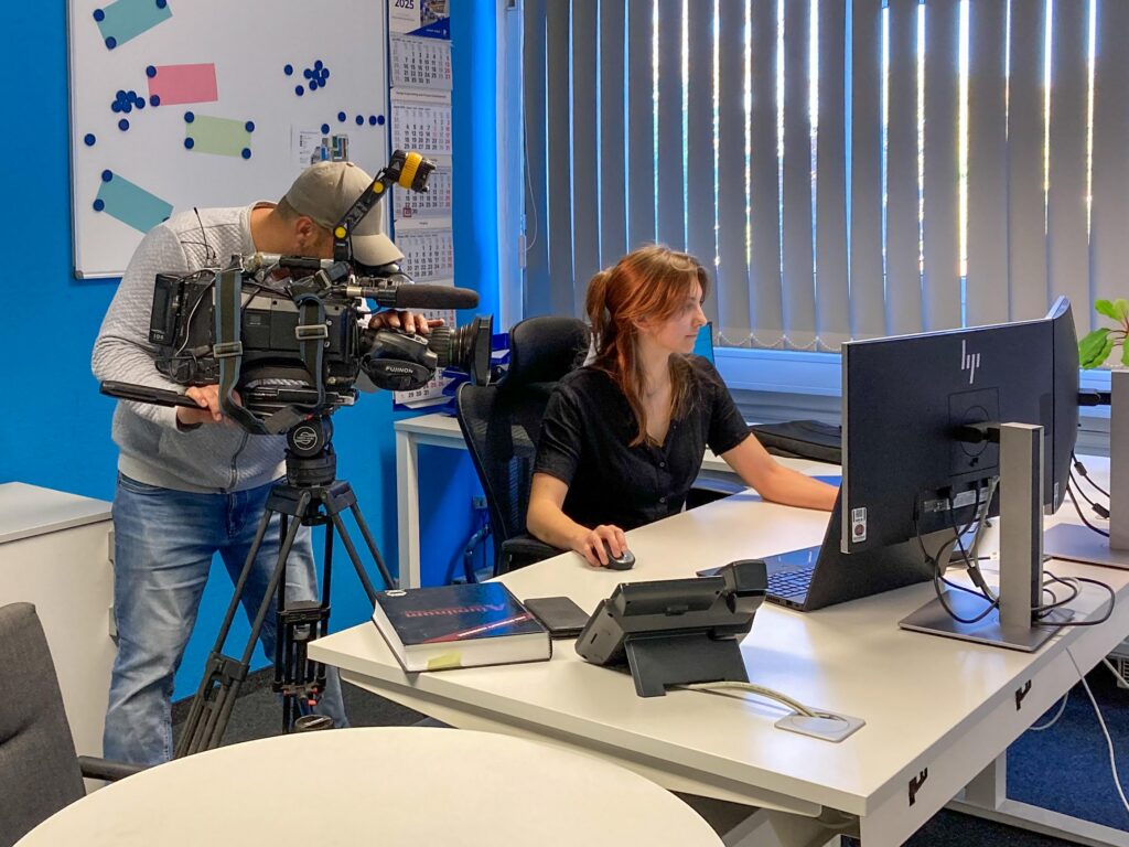 Ein Kameramann filmt eine Frau, die an einem Schreibtisch in einem modernen Büro arbeitet. Die Frau arbeitet an einem Computer, und auf dem Schreibtisch liegen Bücher, ein Telefon und Büromaterial. Vertikale Jalousien verdecken das Fenster hinter ihr.