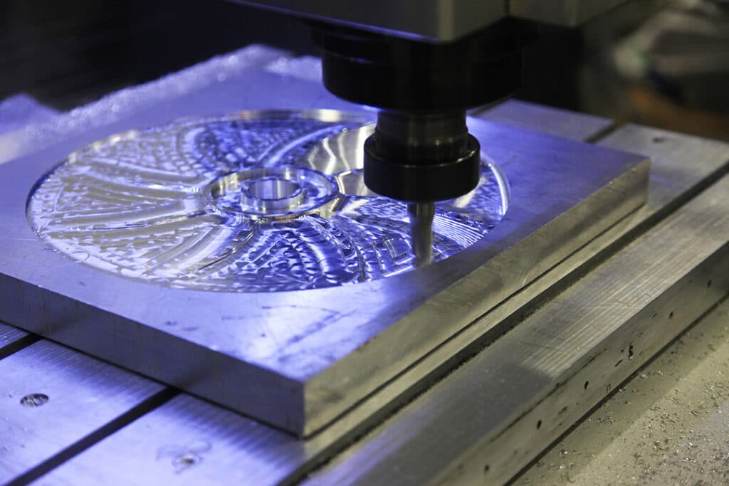 A CNC milling machine cuts a detailed circular pattern into a flat metal plate, illuminated by blue-tinted light inside a workshop.