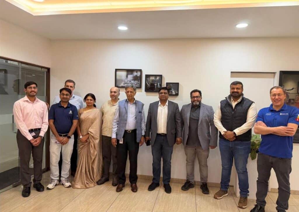 Eine Gruppe von elf Personen, darunter Männer und eine Frau in einem Saree, steht in einem Büro zusammen und lächelt in die Kamera. Gerahmte Fotos hängen an der weißen Wand hinter ihnen.