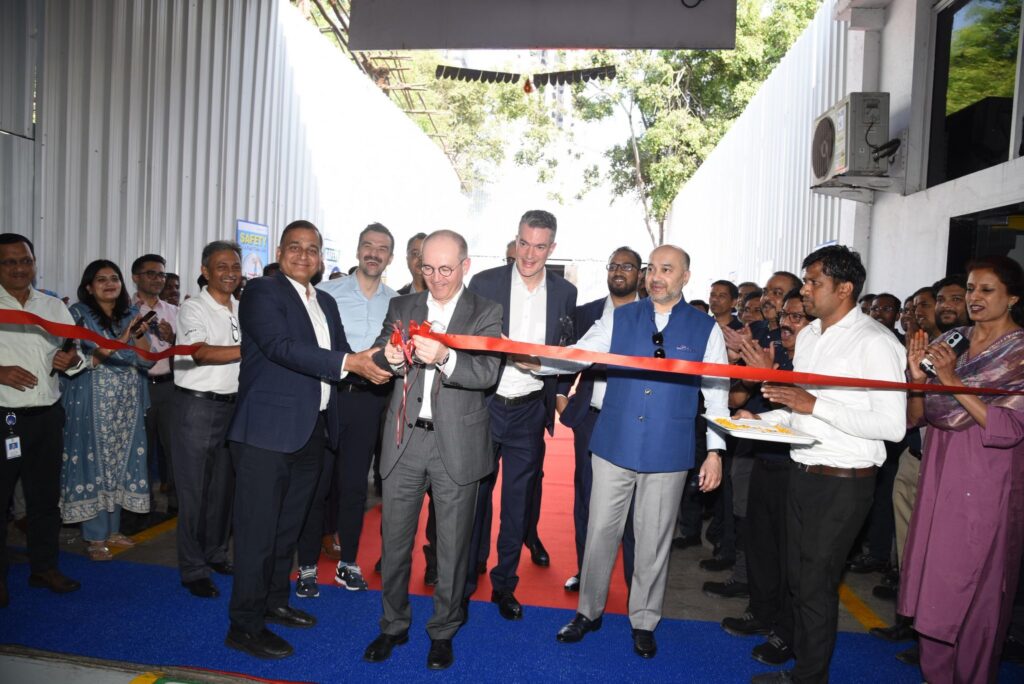 Eine Gruppe von Männern in Geschäftskleidung durchschneidet ein rotes Band bei einer Veranstaltung, umgeben von lächelnden Menschen und Schaulustigen, in einem Außenbereich mit einem blauen Teppich und weißen Wänden.