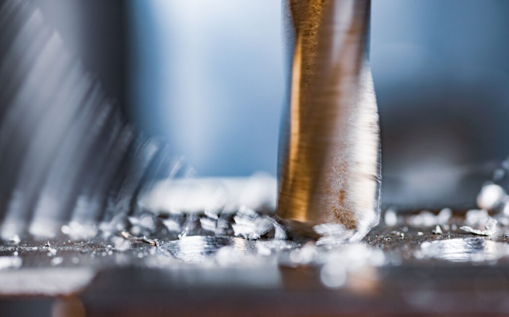 Close-up of a metal drill bit spinning rapidly as it drills into a surface, creating metal shavings and debris around the point of contact.
