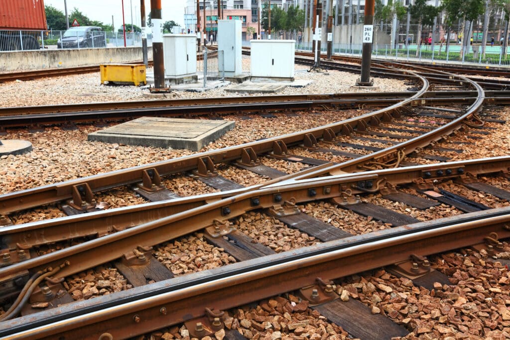Eine Nahaufnahme von sich kreuzenden Eisenbahnschienen mit Schotter dazwischen, umgeben von Stromkästen und städtischen Gebäuden im Hintergrund.