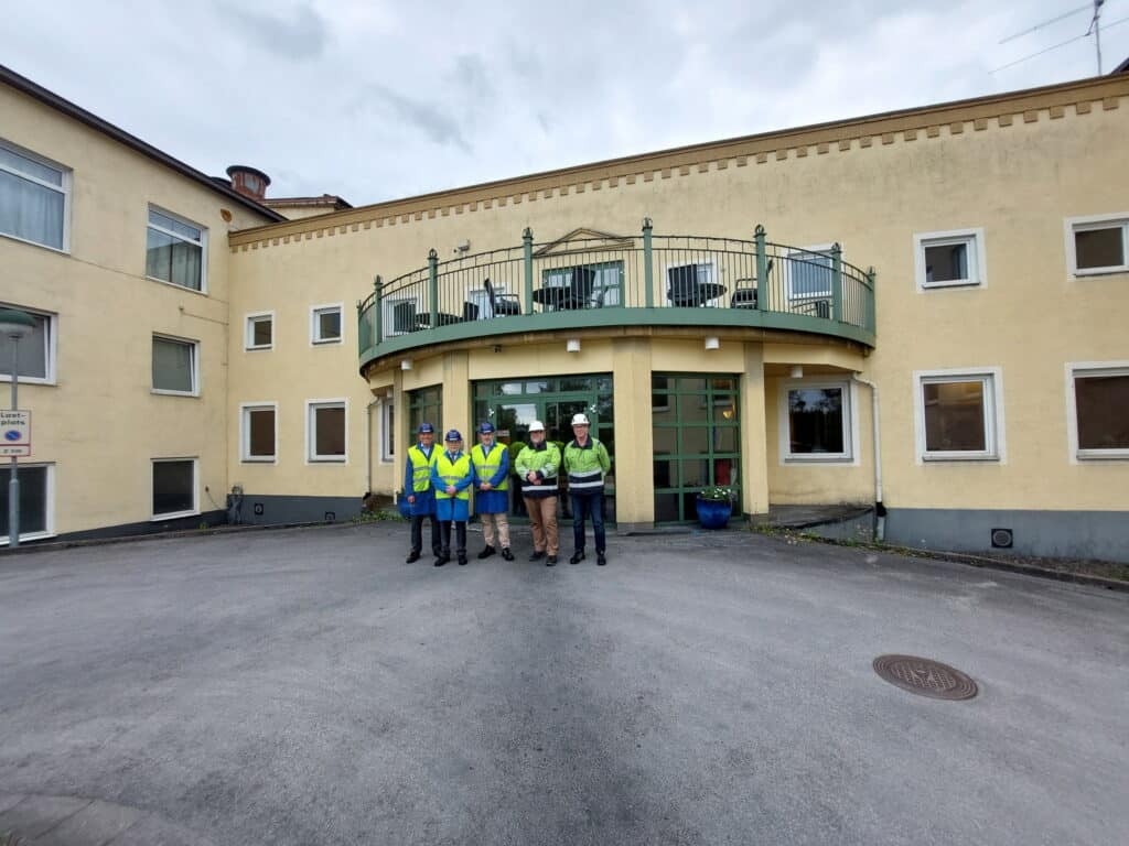 Fünf Personen in Sicherheitswesten und Helmen stehen vor einem gelben Gebäude mit großen Fenstern und einem Balkon über dem Eingang. Die Gruppe befindet sich in der Mitte vor dem Gebäude auf einer gepflasterten Fläche.
