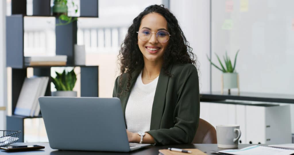 Eine Frau mit lockigem Haar und Brille, die einen dunkelgrünen Blazer trägt, sitzt lächelnd an einem Schreibtisch mit einem Laptop. Im Hintergrund sind Pflanzen und Büromaterial zu sehen.